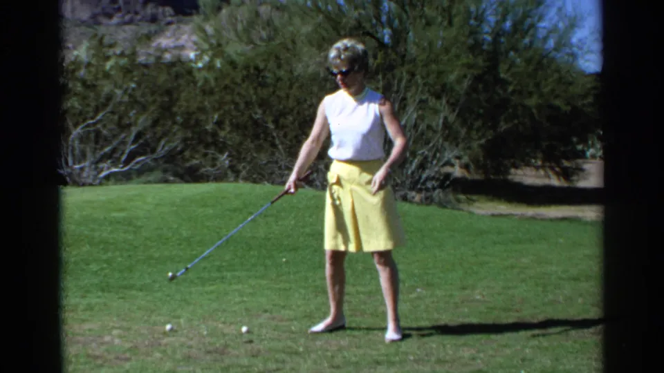 A woman in a yellow skirt holds a golf club on a Scottsdale lawn, framed by shrubs and strong sunlight.