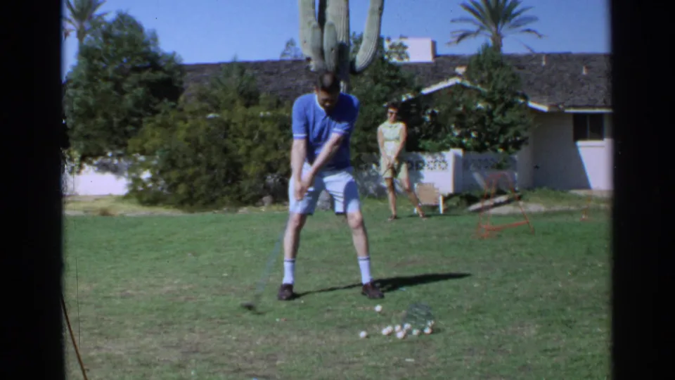 A man in a blue shirt practices golf in Scottsdale, with a woman watching near a saguaro cactus and suburban house.