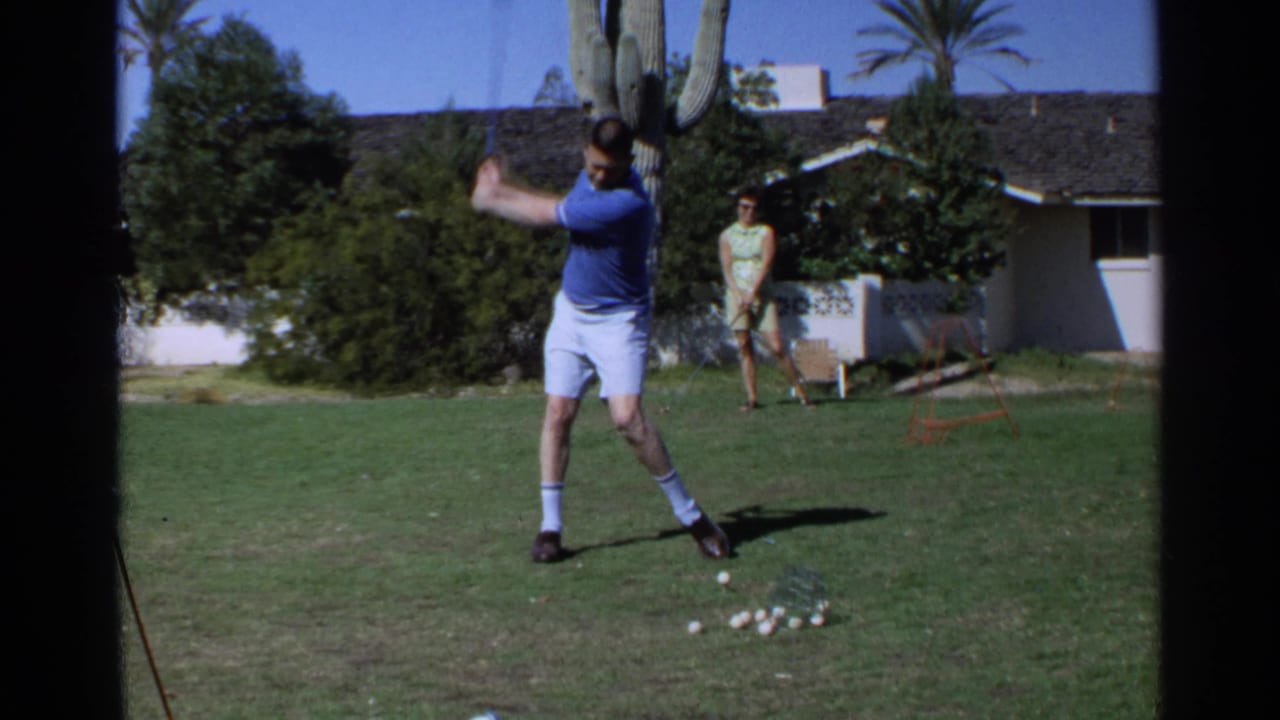 Man in blue shirt swings a golf club on a sunny lawn while a woman stands behind him.