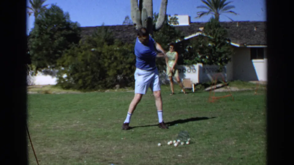The man follows through during a backyard golf practice swing in the 1968 Scottsdale home movie, with golf balls on the lawn and a woman behind him.