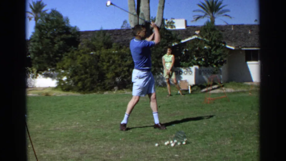 A man holds his golf club high after swinging in a Scottsdale backyard, with a woman standing near shrubs and a low house in the background.