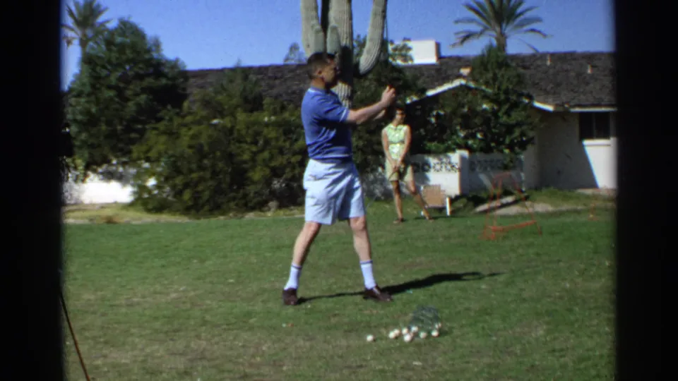 At the end of the 1968 Scottsdale backyard golf shot, the man stands on the lawn after swinging as a woman watches near the house and cactus.