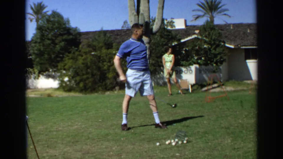 A man in a blue shirt follows through on a backyard golf swing in Scottsdale, Arizona, with a woman, house, cactus, and palm trees behind him.