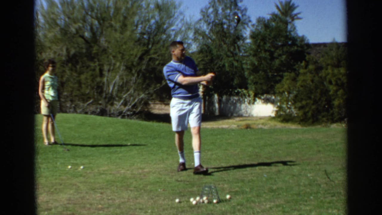 Man in blue polo swings a golf club on a lawn while a woman stands nearby.