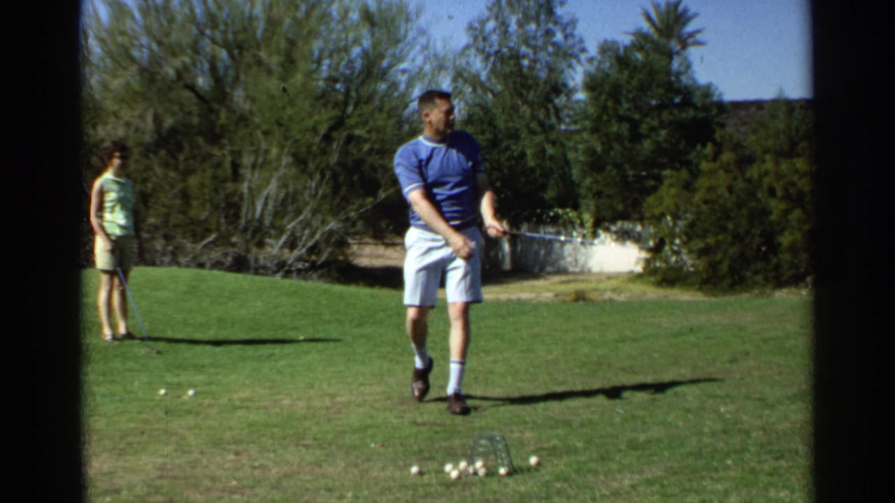 Man walks after a golf swing with a woman watching from the lawn behind him.