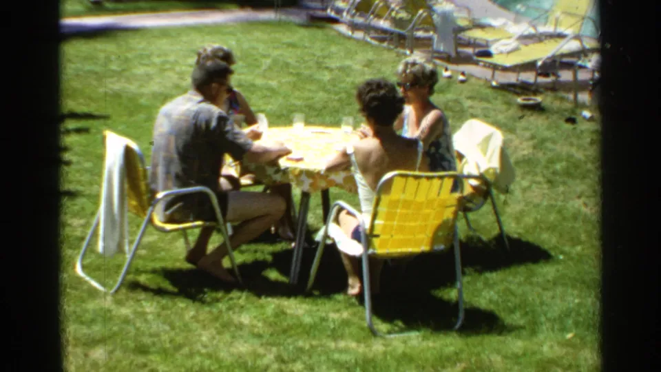 1968 Scottsdale home-movie frame of four adults dining outdoors at a table with yellow chairs and poolside furniture nearby.