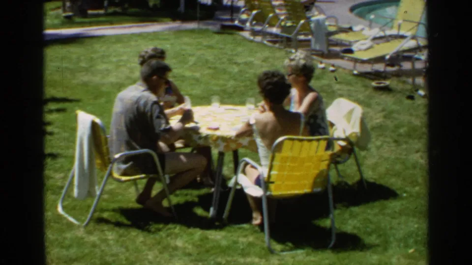 1968 Scottsdale home-movie frame showing adults at a lawn table beside yellow patio seating, with poolside lounge furniture in the background.