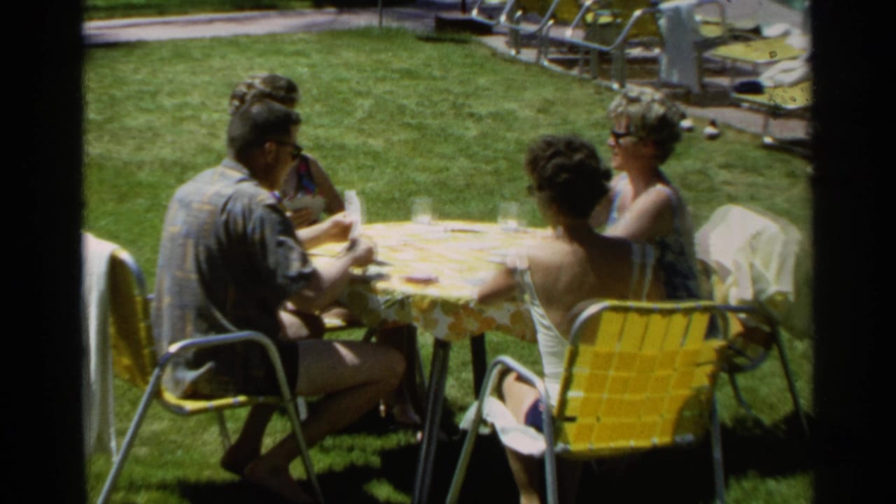 Four adults sit around a small outdoor table on a lawn, with cards and glasses visible.