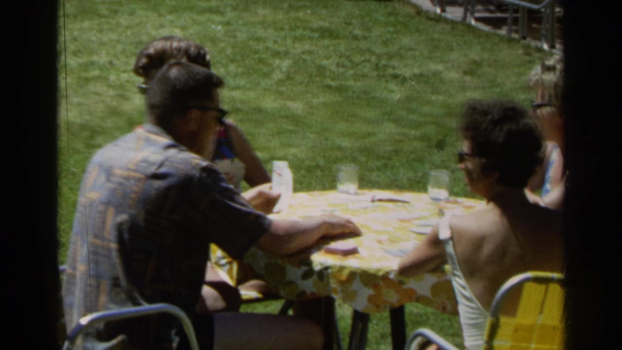 Adults in sunglasses sit at a backyard table with cards and glasses on the grass.