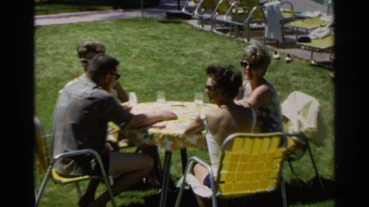 Four adults continue the backyard card game in Scottsdale, with yellow lawn chairs, glasses, and poolside furniture visible.