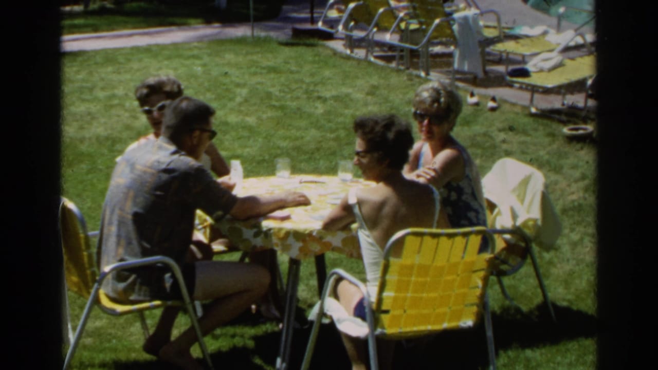 1968 Scottsdale home-movie frame of adults gathered at a lawn picnic table with yellow chairs and pool loungers nearby.