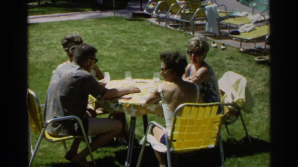 1968 Scottsdale home-movie frame of adults gathered at a lawn picnic table with yellow chairs and pool loungers nearby.
