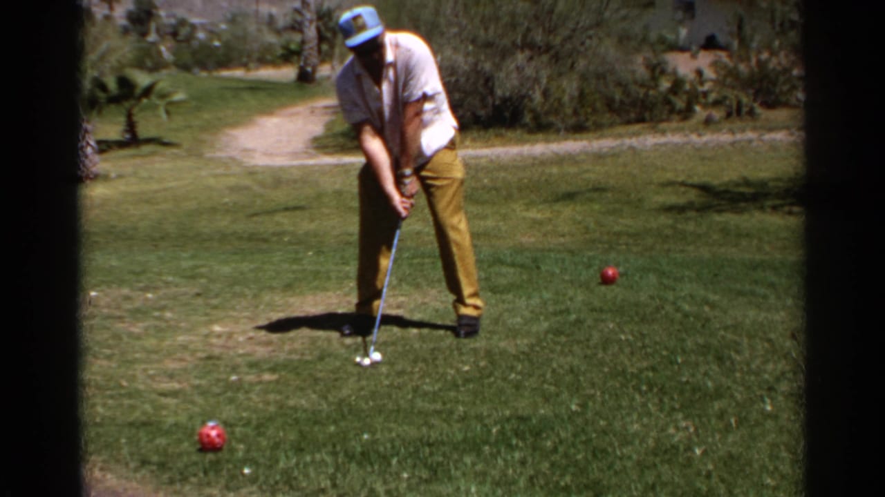 Man addresses a white golf ball on grass with two red balls nearby.