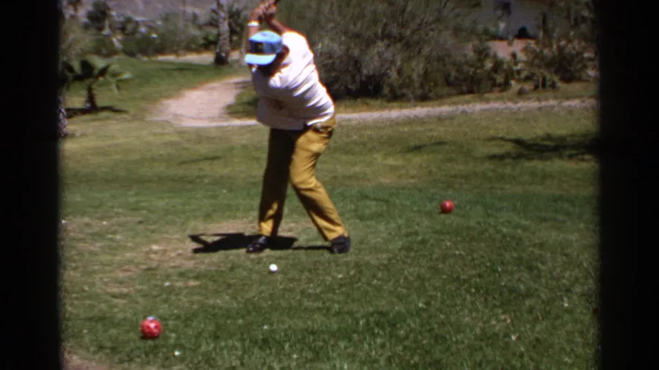 A man in vintage golf attire turns through his swing on the Scottsdale course, framed by grass, shrubs, and a pale dirt path.
