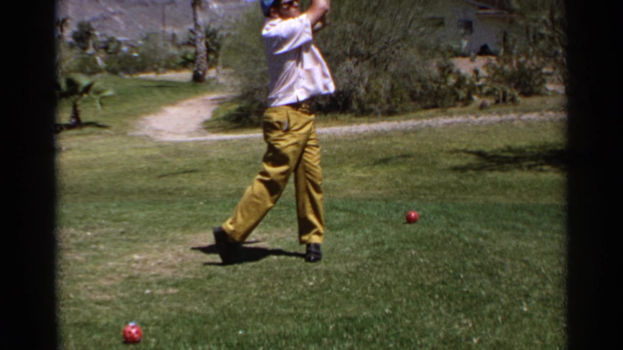 Man in a white shirt and yellow trousers swings a golf club on a sunny grass course.