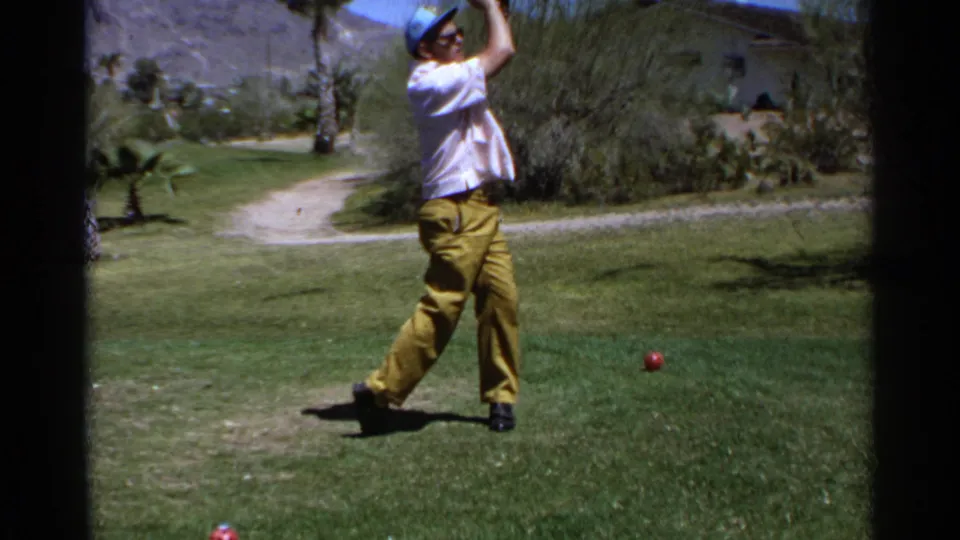 The 1968 home-movie frame shows the golfer mid-finish on a desert course, with grass, a path, shrubs, palms, and mountains visible.