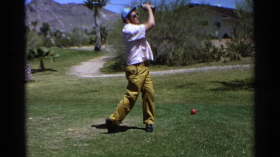 A man in a cap and yellow trousers completes a golf swing in supported 1968 Scottsdale footage, framed by palms, shrubs, and distant mountains.