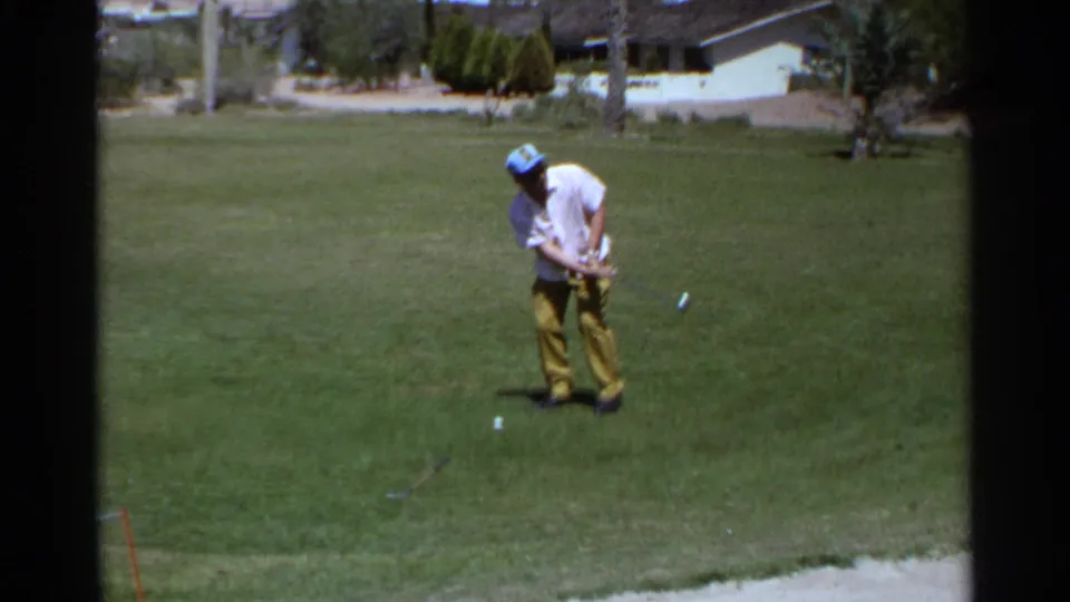 Young man makes a short golf stroke on a Scottsdale course, framed by grass, trees, and low buildings in the distance.