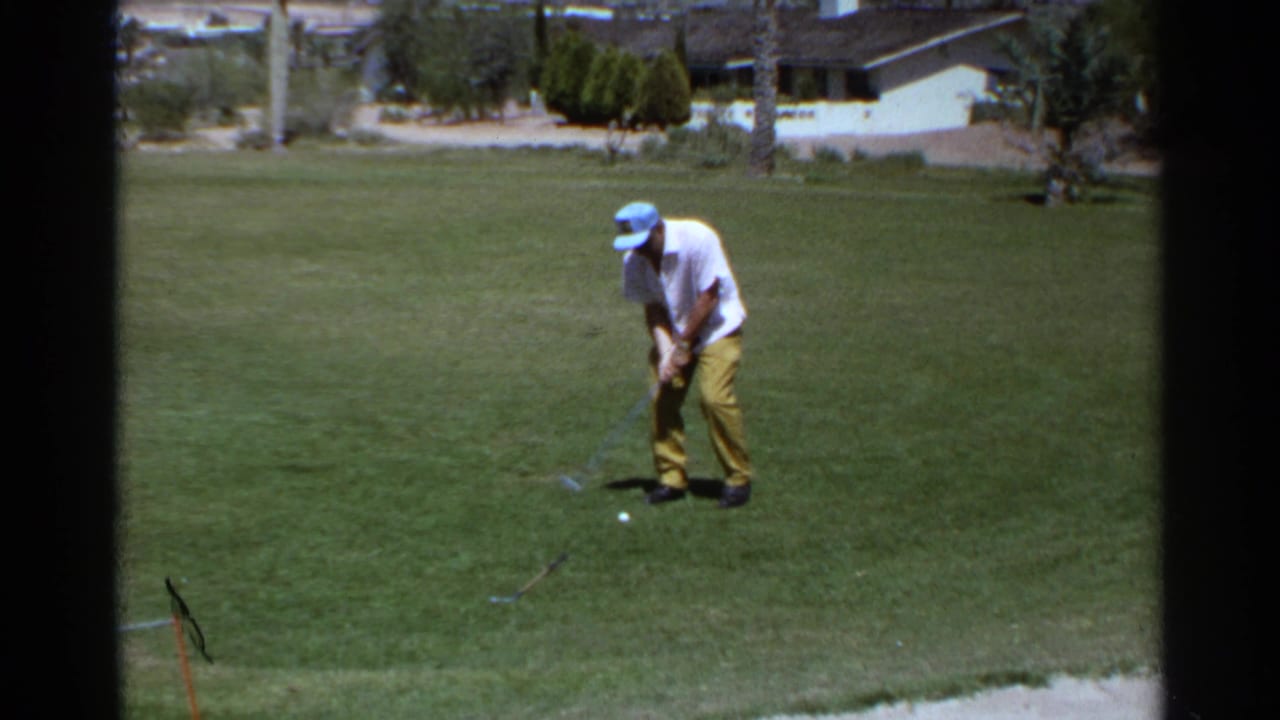 Adult golfer practices on a Scottsdale, Arizona green in 1968, with a sand trap edge, palms, and low houses behind him.