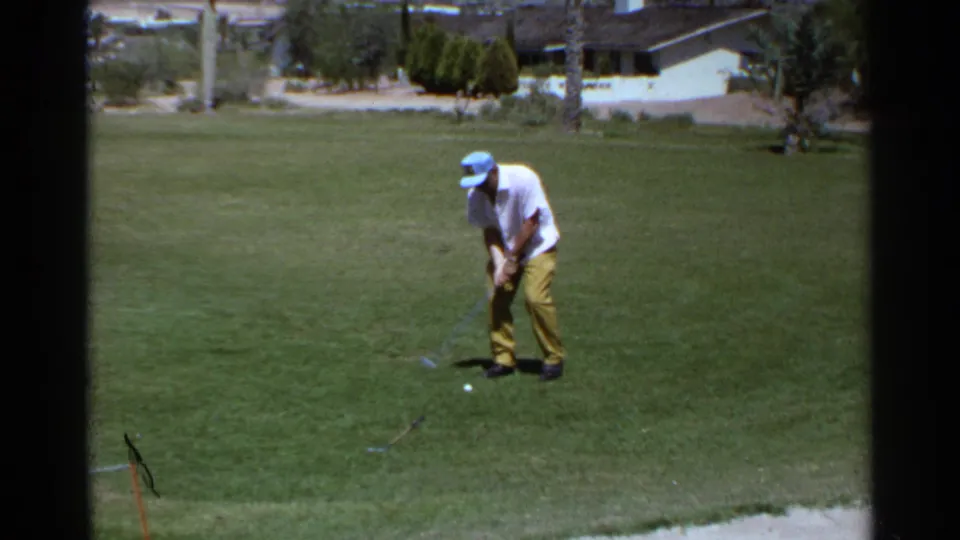 Adult golfer practices on a Scottsdale, Arizona green in 1968, with a sand trap edge, palms, and low houses behind him.