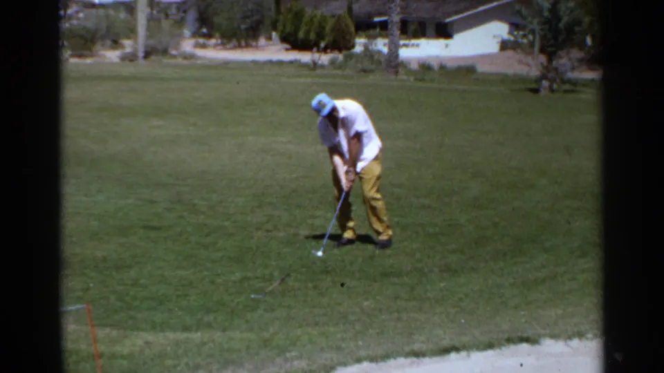 In the 1968 Scottsdale home-movie frame, the golfer leans into his stance on a sunlit green near a pale sand trap edge.