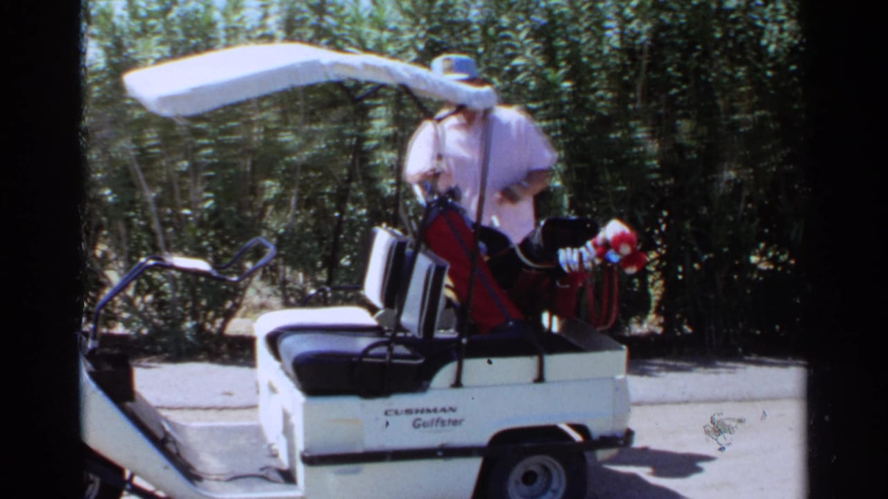 A man sits in a white Cushman golf cart with golf clubs beside trees during the 1968 Scottsdale golf course footage.
