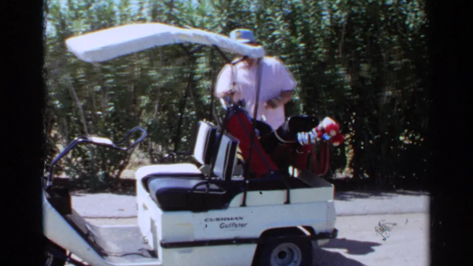A man sits in a white Cushman golf cart with golf clubs beside trees during the 1968 Scottsdale golf course footage.