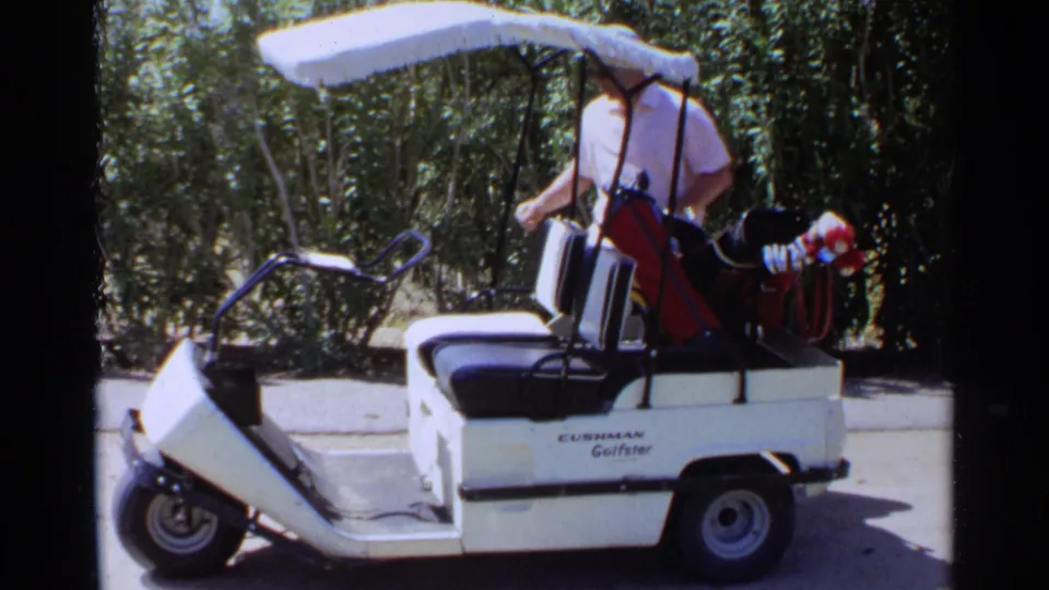 In Scottsdale, Arizona in 1968, a man stands by a white Cushman Gulfster golf cart with clubs on a sunny path.