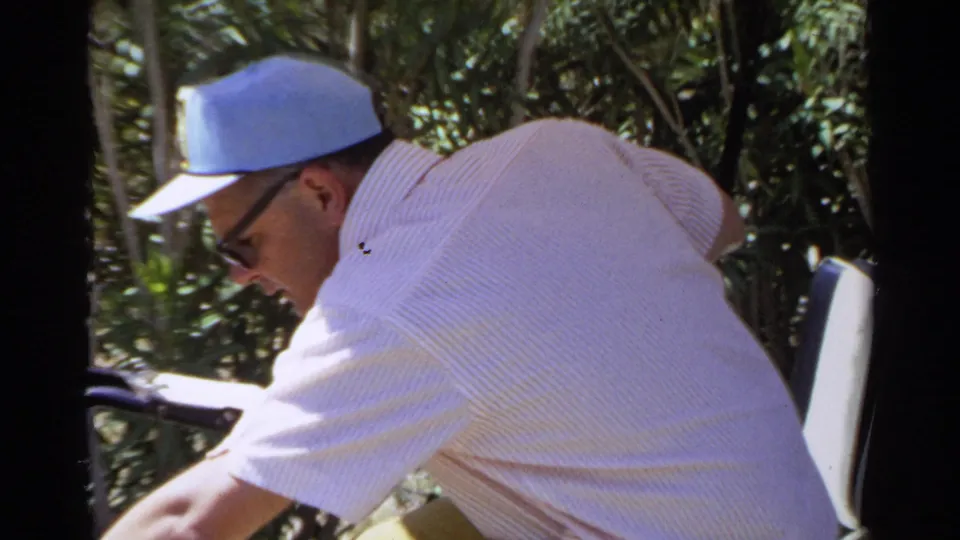 The man leans over the front of the golf cart, with his blue cap, sunglasses, light shirt, and leafy background visible.