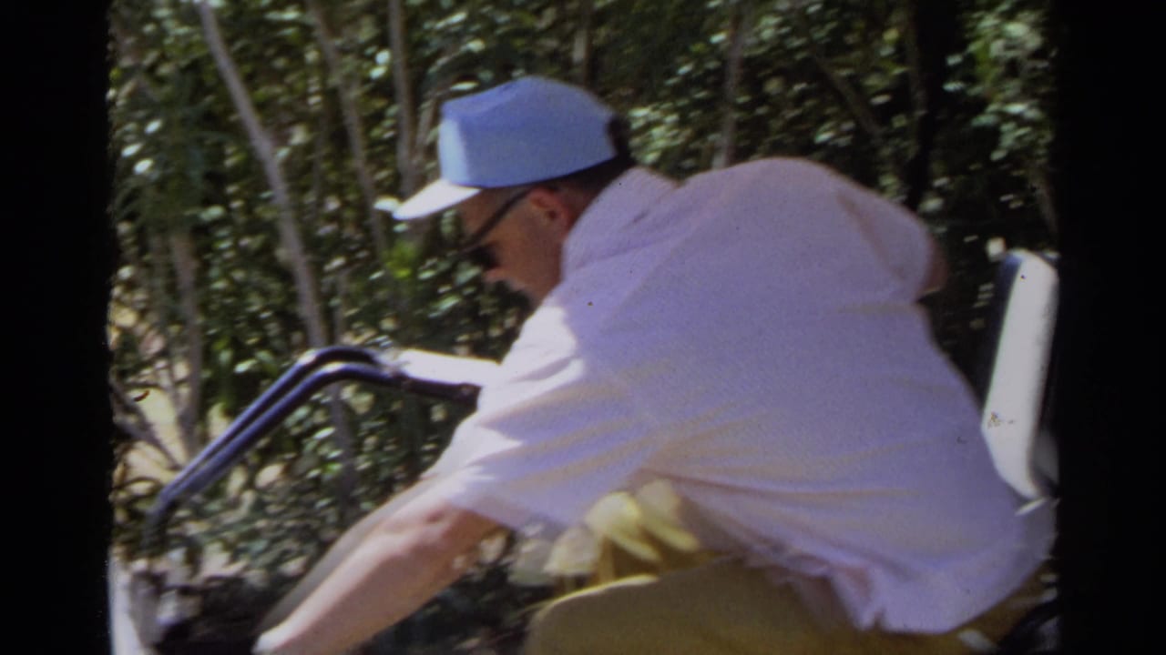 Older man in a light blue cap rides a golf cart beside trees and shrubs.