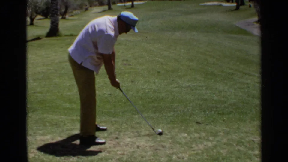 Older man in cap and sunglasses sets up a golf shot on a sunlit Scottsdale course with trees and grass visible.