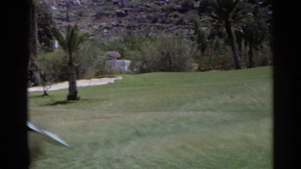 Vintage 8mm view of a Scottsdale fairway with palms, sand trap, shrubs, and rocky hillside beyond.