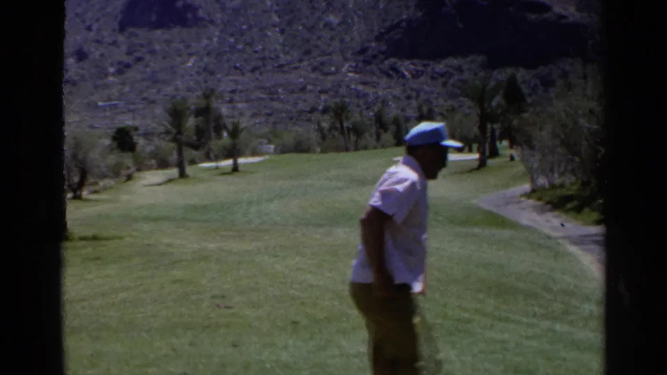 A man in a light shirt and blue cap walks across a Scottsdale golf fairway holding a club, with palms and hillside behind him.