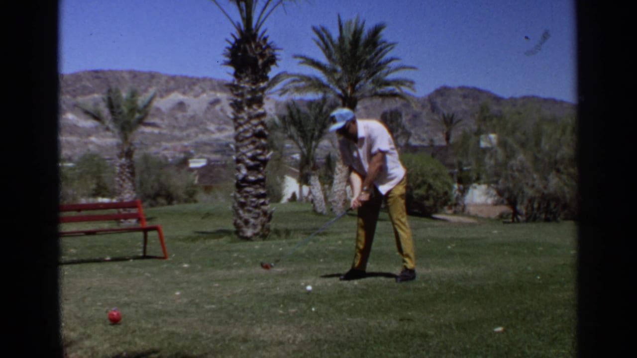 Adult golfer in a white shirt and yellow pants prepares a shot on a Scottsdale golf course with palms and desert hills behind.