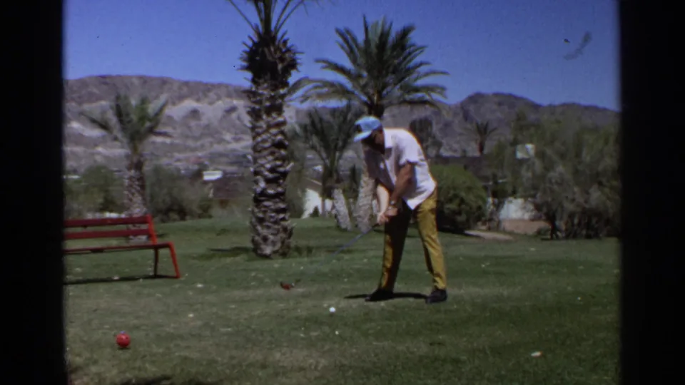 Adult golfer in a white shirt and yellow pants prepares a shot on a Scottsdale golf course with palms and desert hills behind.