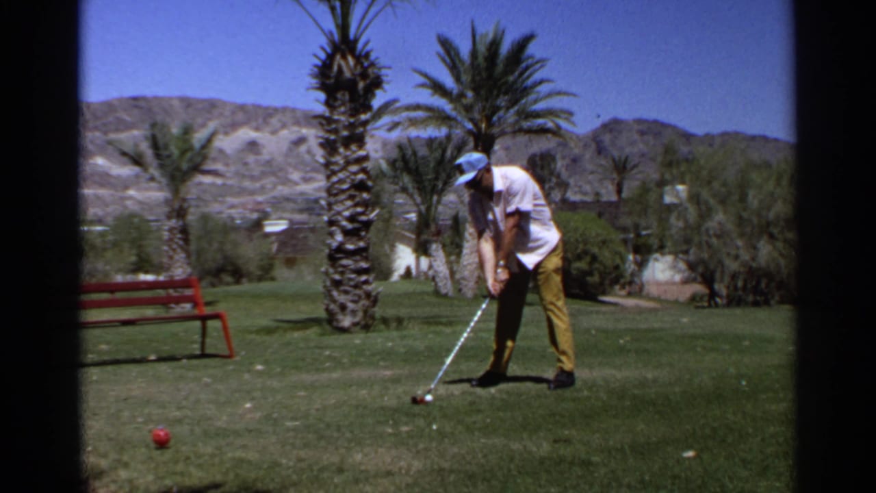 1968 Scottsdale frame of an adult golfer addressing the ball on a green course bordered by palms, a red bench, and desert hills.