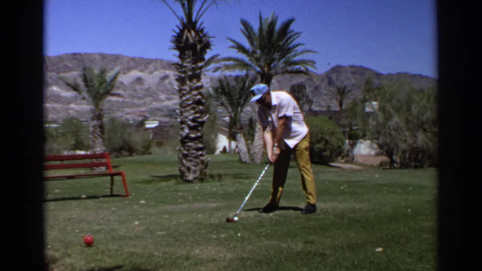 1968 Scottsdale frame of an adult golfer addressing the ball on a green course bordered by palms, a red bench, and desert hills.