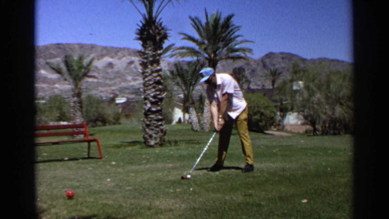 In Scottsdale, Arizona in 1968, an adult golfer in a white shirt, yellow pants, and blue cap lines up a shot on a grassy area near palms.