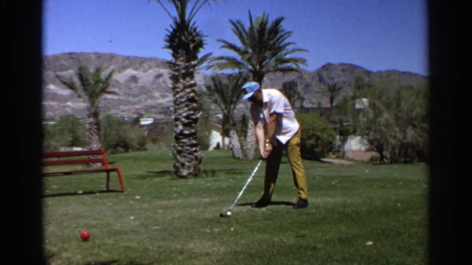 In Scottsdale, Arizona in 1968, an adult golfer in a white shirt, yellow pants, and blue cap lines up a shot on a grassy area near palms.