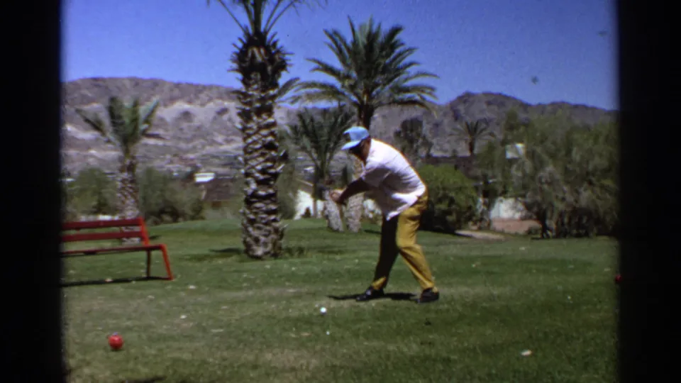 In this Scottsdale 1968 frame, the golfer rotates into his swing while a red bench, palm trees, lawn, and desert hills remain visible.