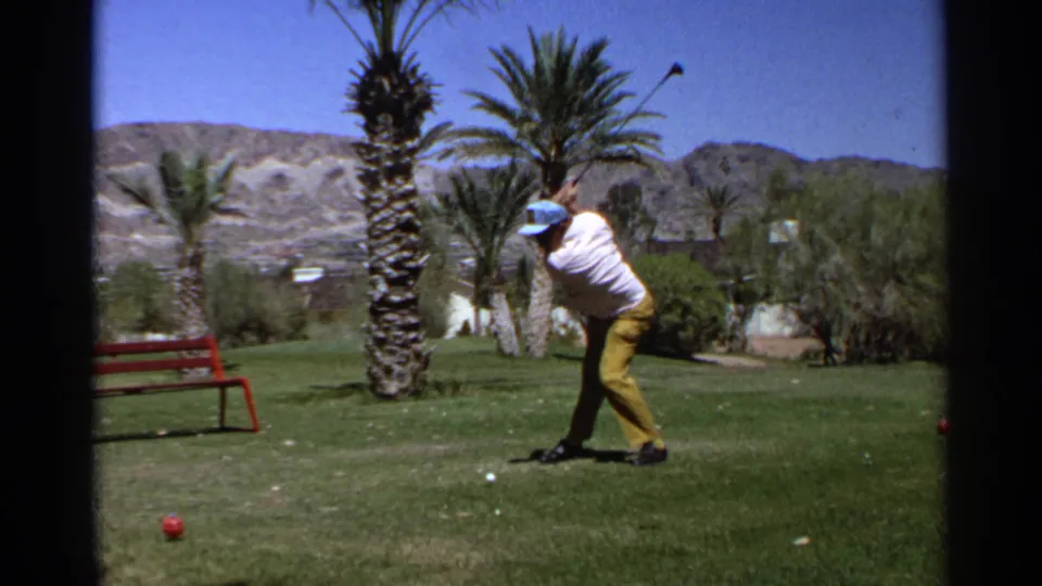 The golfer follows through in the 1968 Scottsdale scene, his club raised as palms, grass, a red bench, and desert mountains fill the frame.