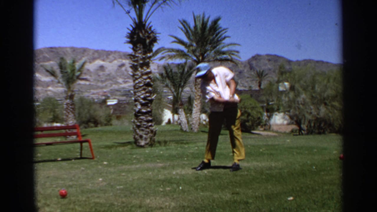 Adult golfer bends forward on grass with a club near palm trees and a red bench.