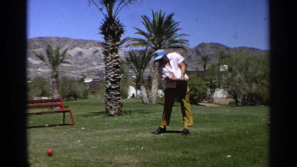 In a 1968 Scottsdale, Arizona 8mm frame, an adult golfer in a light cap, white shirt, and yellow trousers bends over on a grassy desert course with palm trees, a red bench, and hills behind him.