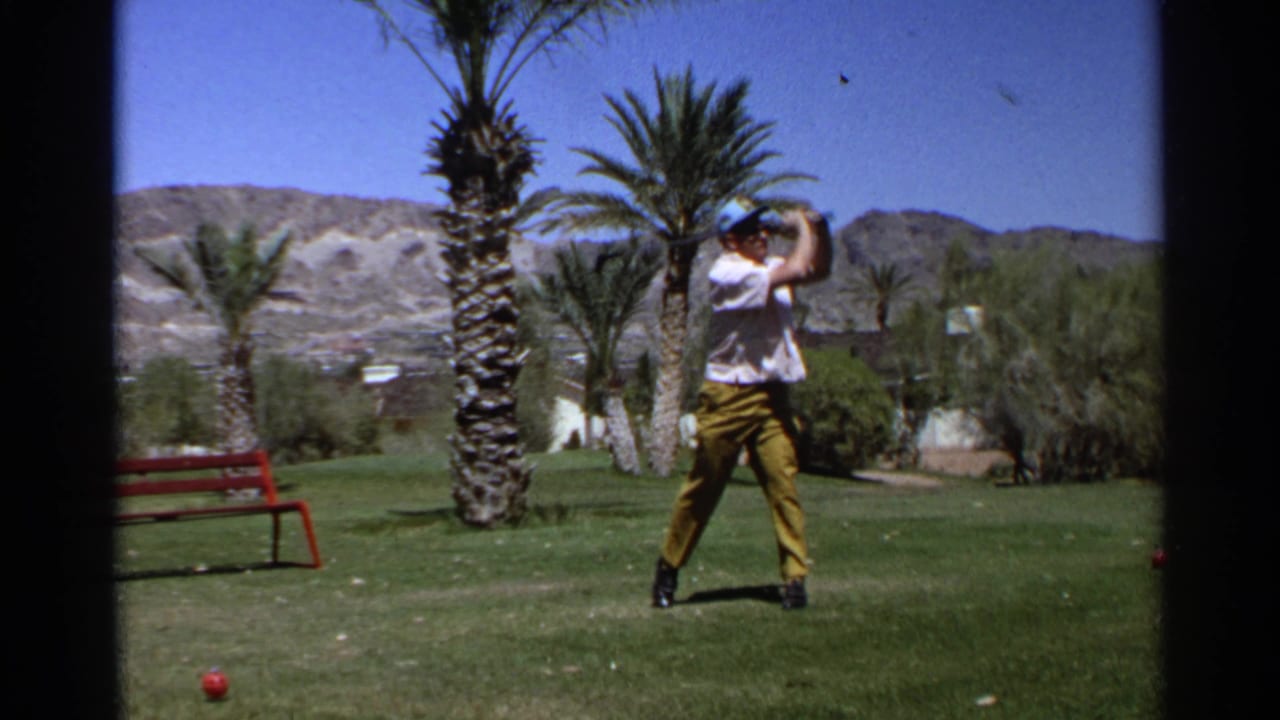 Adult golfer raises a club during a swing on a palm-lined course.