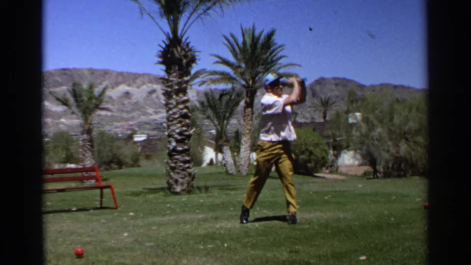 The golfer raises the club high during his swing on the Scottsdale grass course, with palm trunks, desert shrubs, a red bench, and mountains visible in the background.