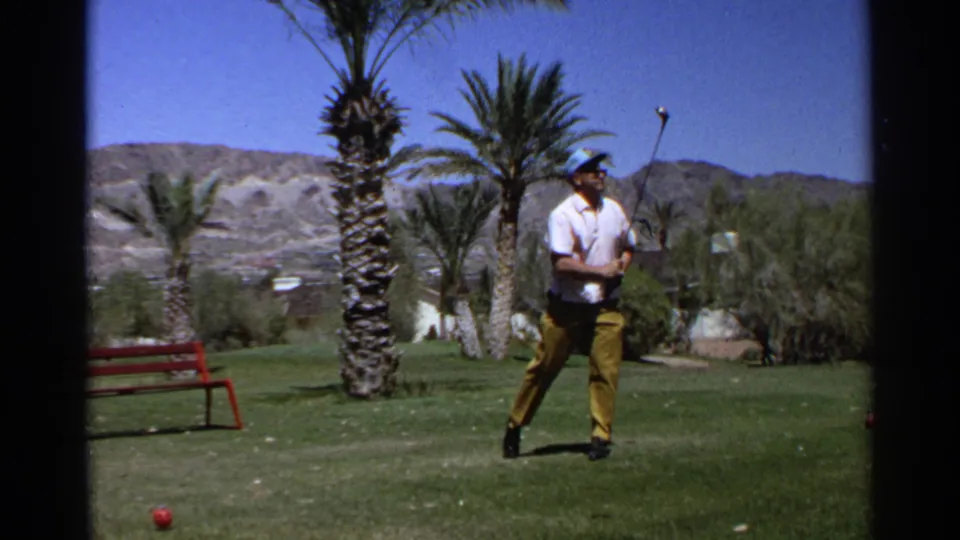 At the end of the 1968 Scottsdale golf shot, the adult golfer stands on the grass with his club raised, with palm trees, desert vegetation, a red bench, and mountains behind him.