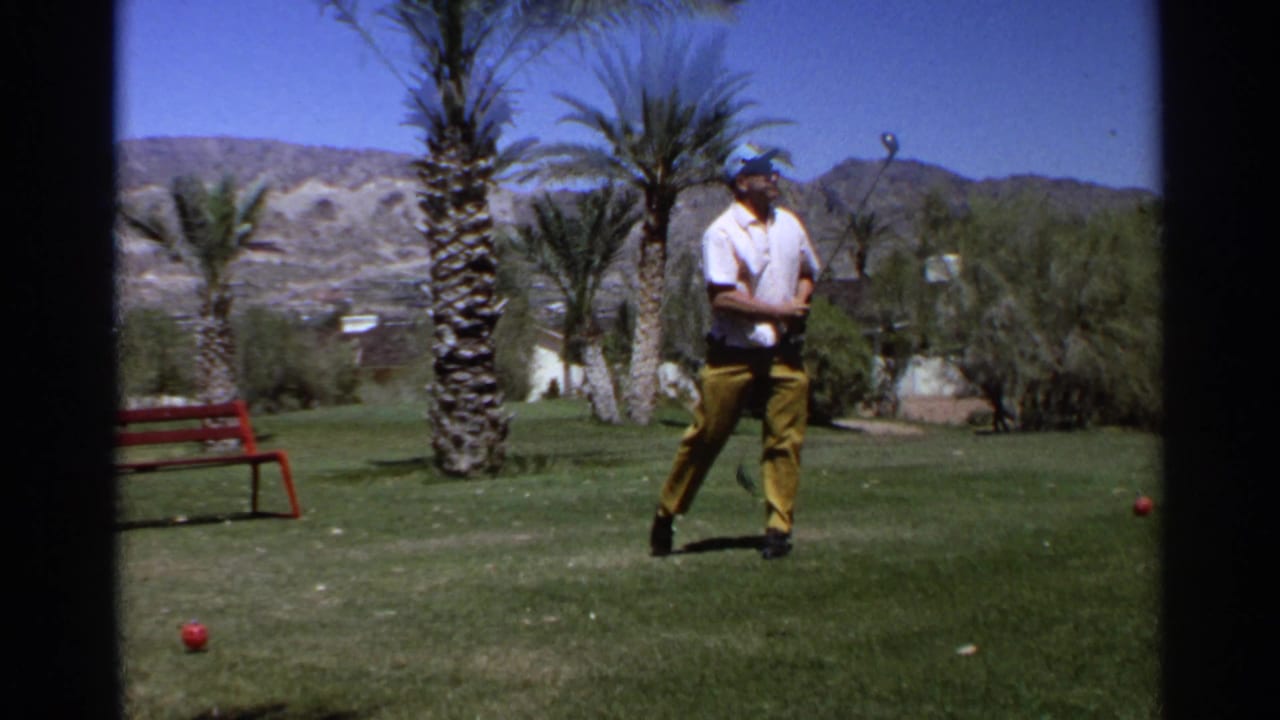 In supported 1968 Scottsdale footage, a man in a white shirt and yellow trousers stands on a golf course with palm trees and mountains behind him.