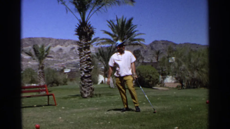 A man in yellow trousers holds a golf club and looks toward the ground on a 1968 Scottsdale golf course bordered by palms and desert hills.