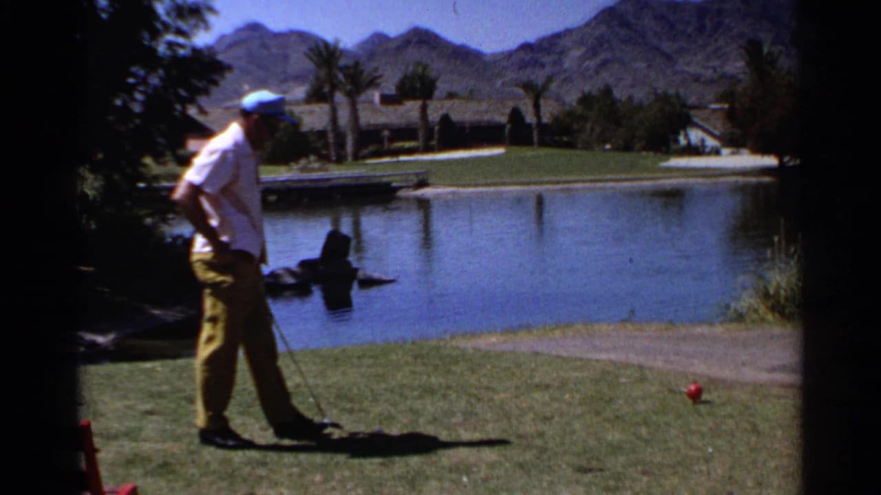 In 1968 Scottsdale, an adult golfer stands near a water hazard with palms, lawn, and desert mountains behind him.