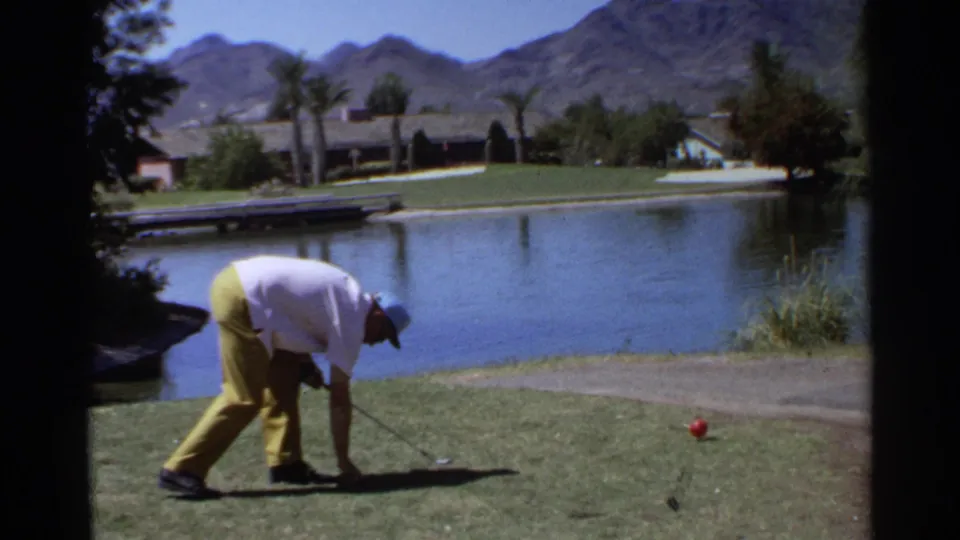 The Scottsdale vacation golfer bends to adjust or retrieve something near the grass, with water, palms, and mountains visible.